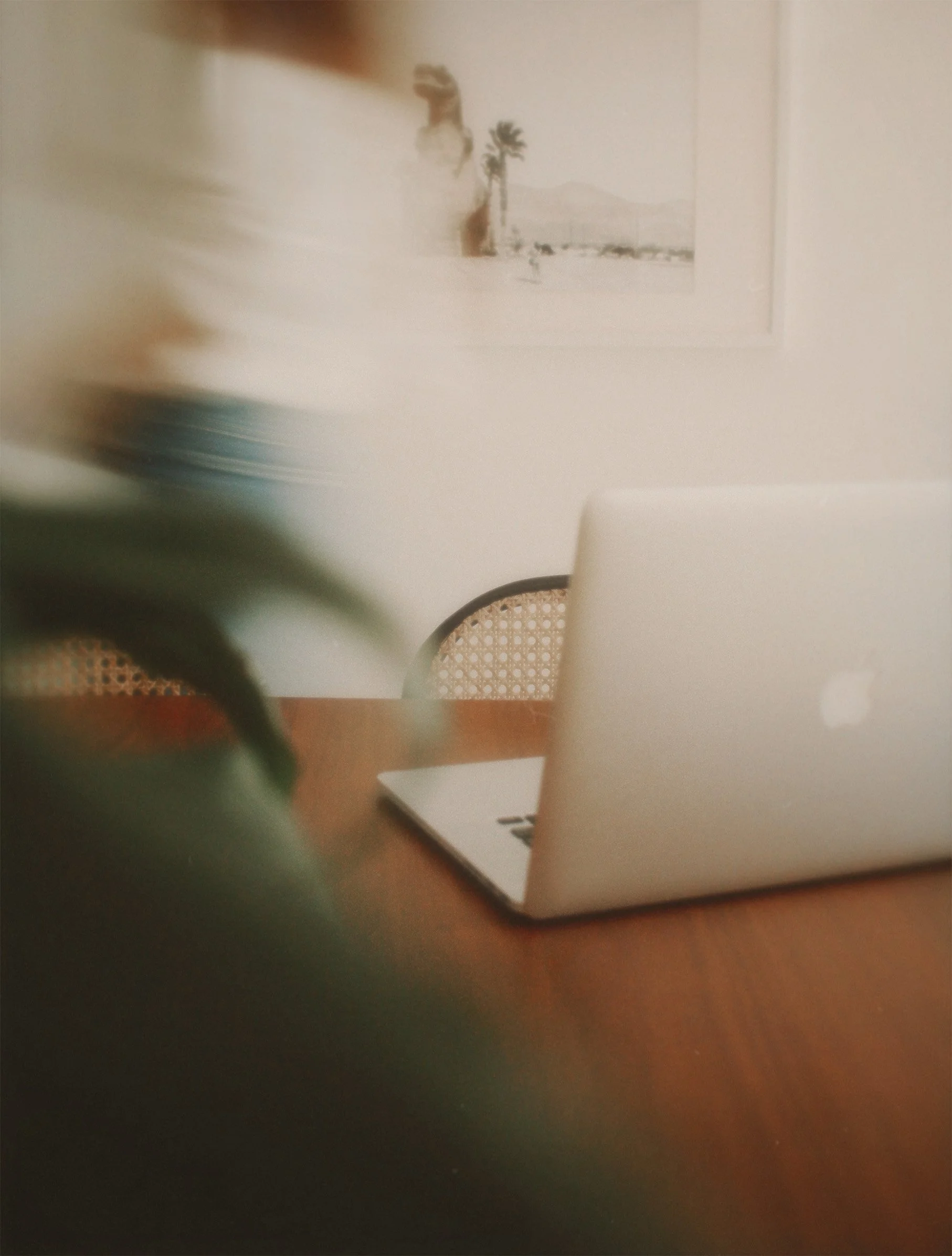 Blurred photo of a wooden table with a silver laptop, a chair in the background, and a framed desert landscape picture hanging on a white wall.