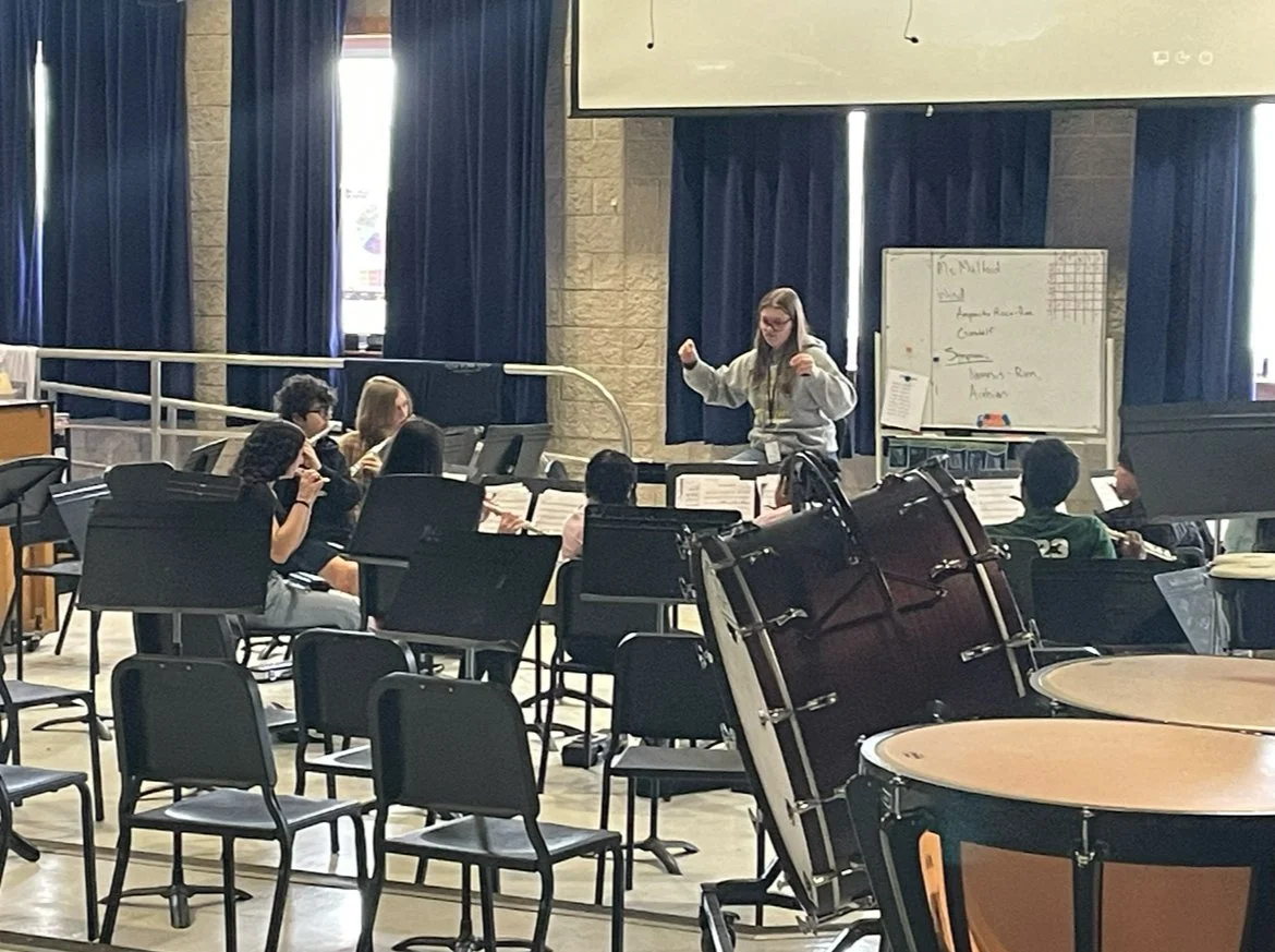 A music rehearsal room with students practicing, a young woman standing and conducting, musical instruments like drums in the foreground, a whiteboard in the background, and chairs arranged around the room.