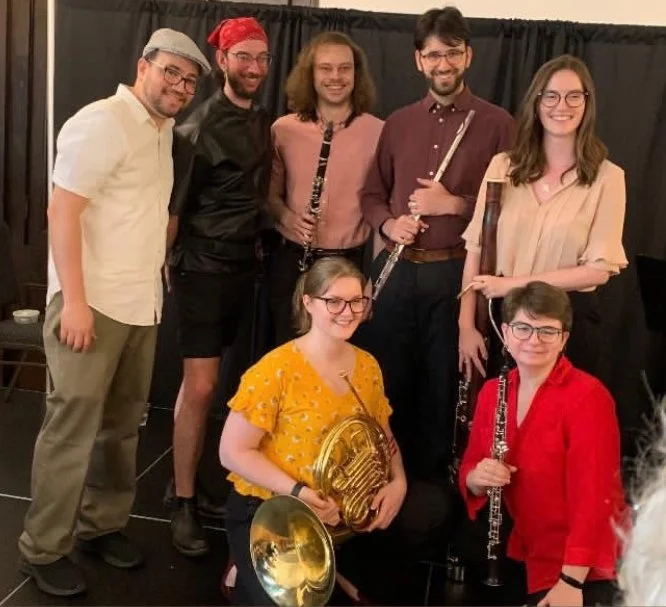 Group of seven people, six standing and one sitting, posing with musical instruments in front of a black curtain, smiling at the camera.