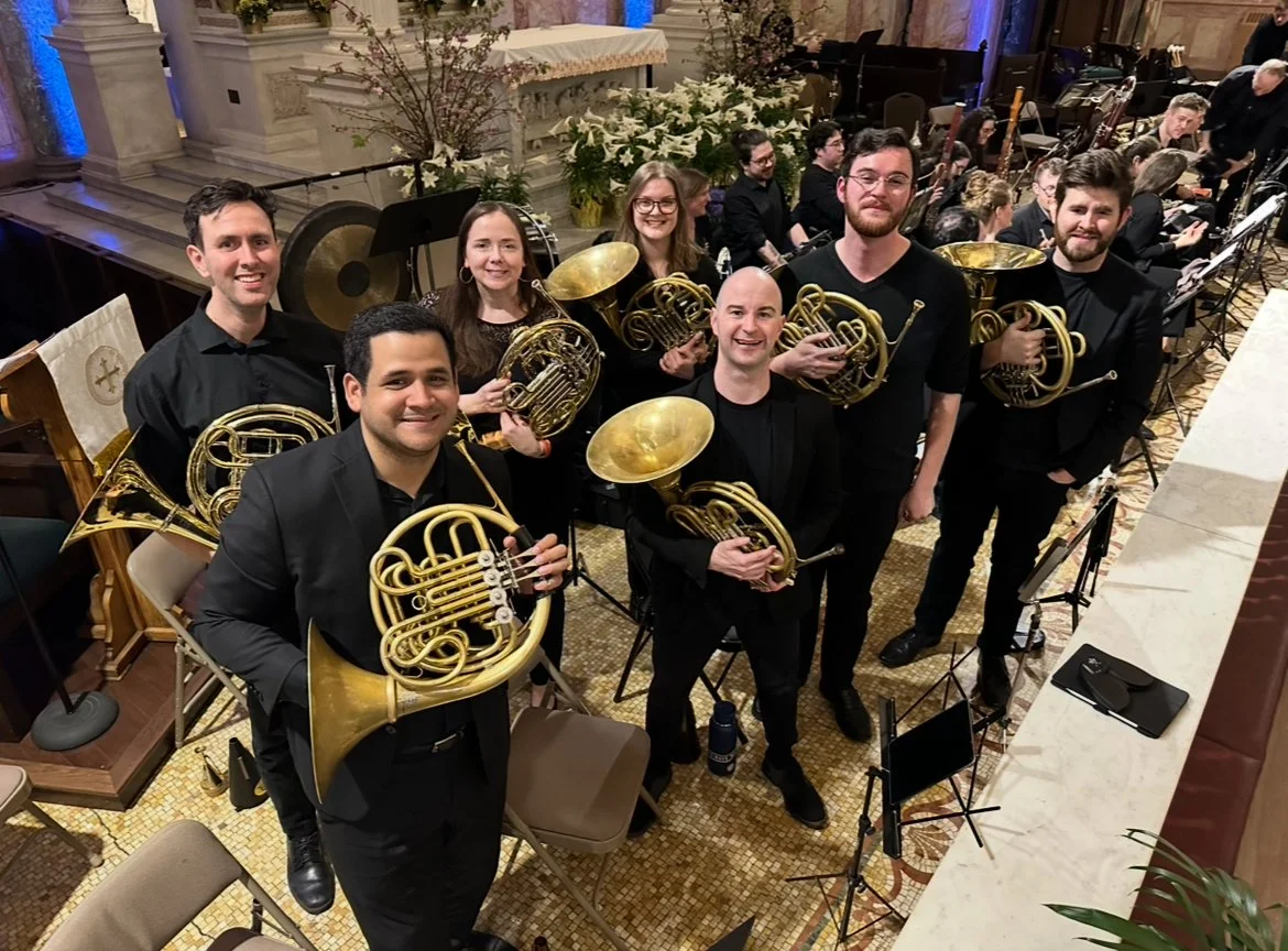 A group of musicians holding French horns and other brass instruments in a concert hall, smiling at the camera.