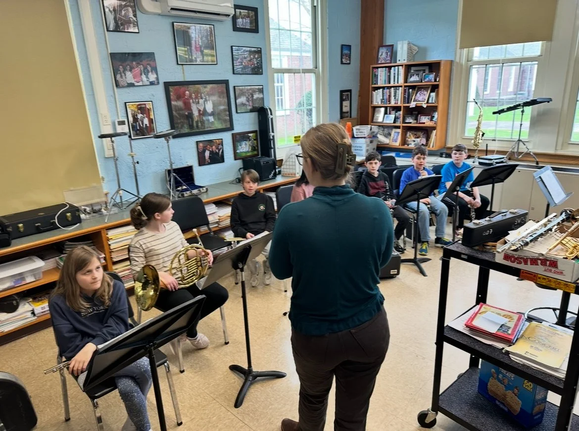 Children practicing music in a classroom with a teacher, musical instruments, and music stands.