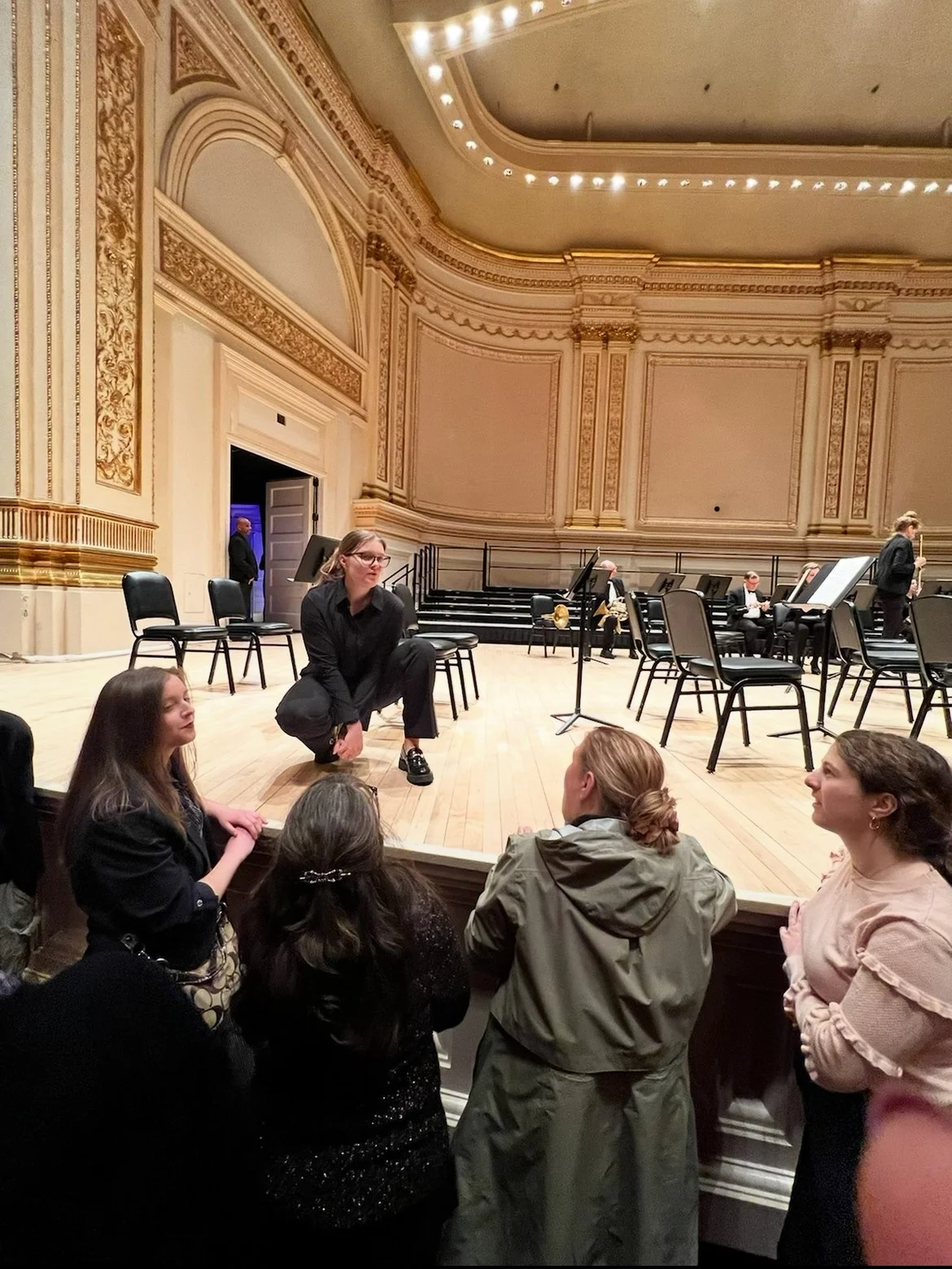 A young woman crouching on stage engaging with a group of four women standing and sitting near the front of the stage in a concert hall. The hall has ornate gold and white decoration.