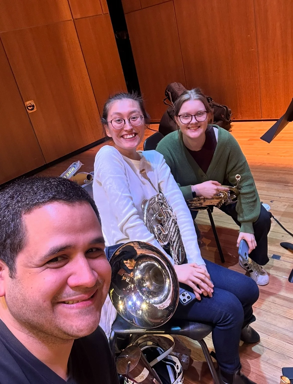 Four people sitting together in a music rehearsal room, holding musical instruments such as a French horn and a trumpet, smiling for a selfie.