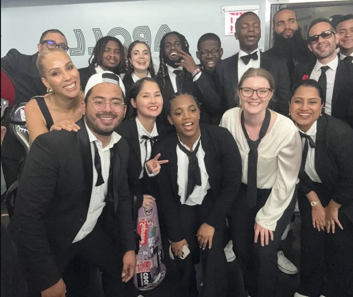 Group of diverse young adults dressed in formal black and white clothing, smiling and posing together in an indoor setting.