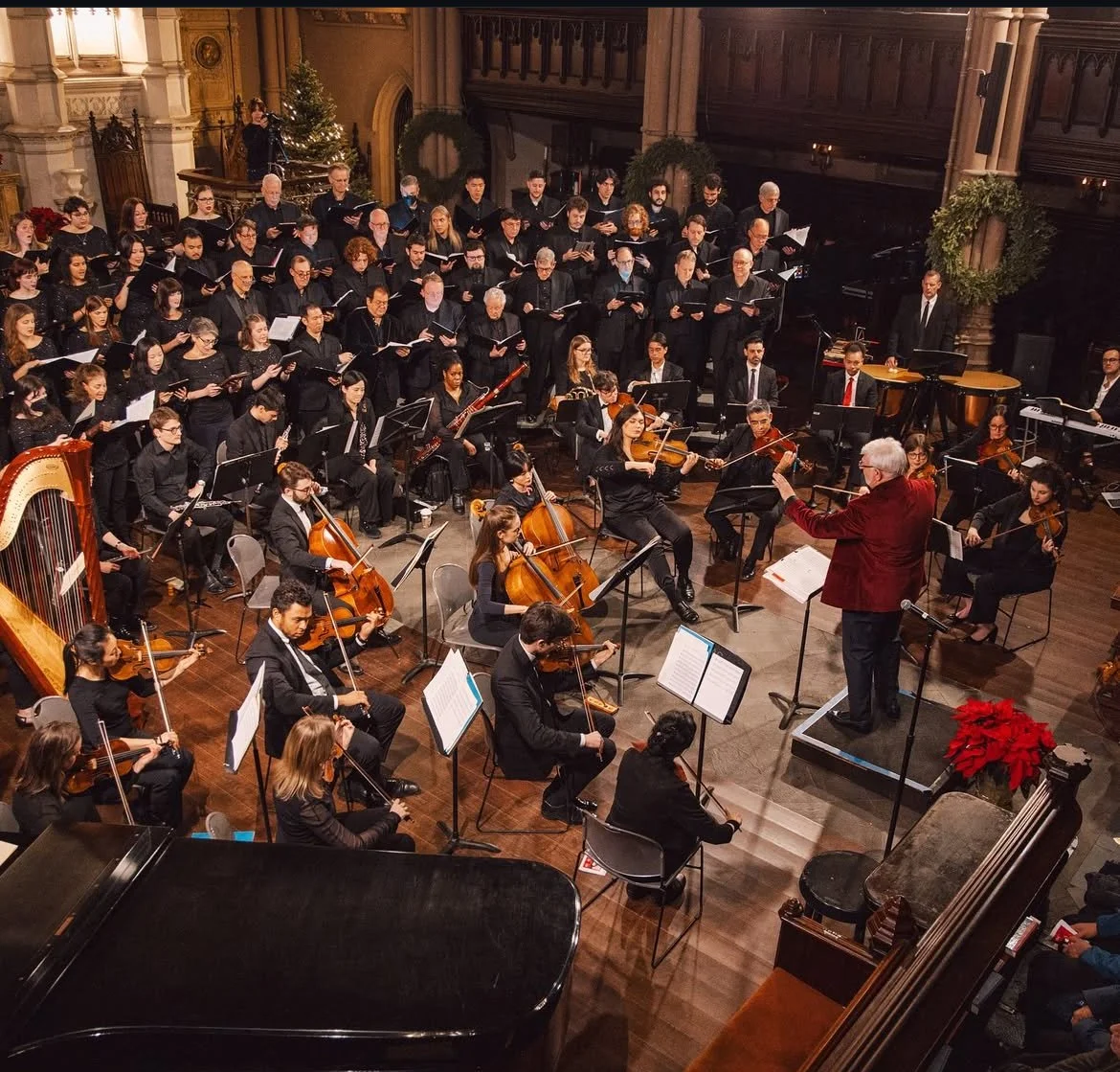 An orchestra performing in a church or concert hall with string, woodwind, and percussion instruments, directed by a conductor in a red jacket.