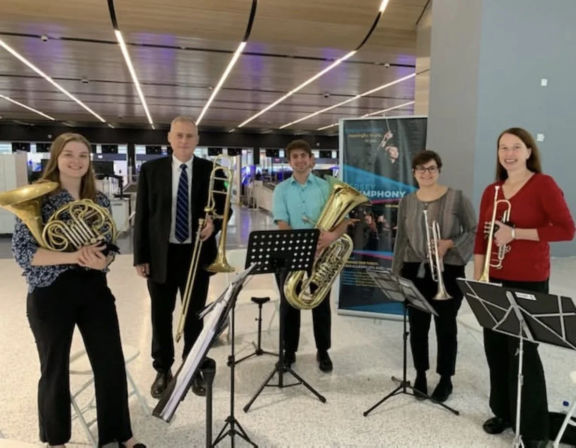 Five musicians holding brass instruments in an airport terminal, with music stands and a poster in the background.
