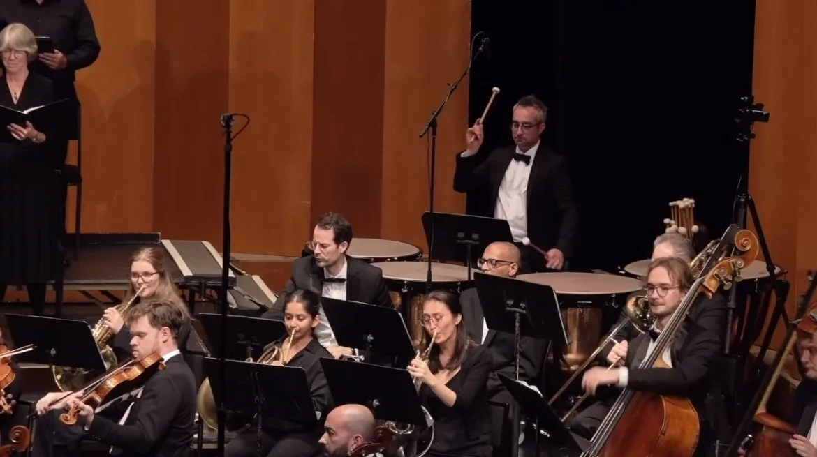 Orchestra performing on stage with musicians playing various instruments, including violin, cello, flute, and percussion, in a concert hall with wooden panel walls.
