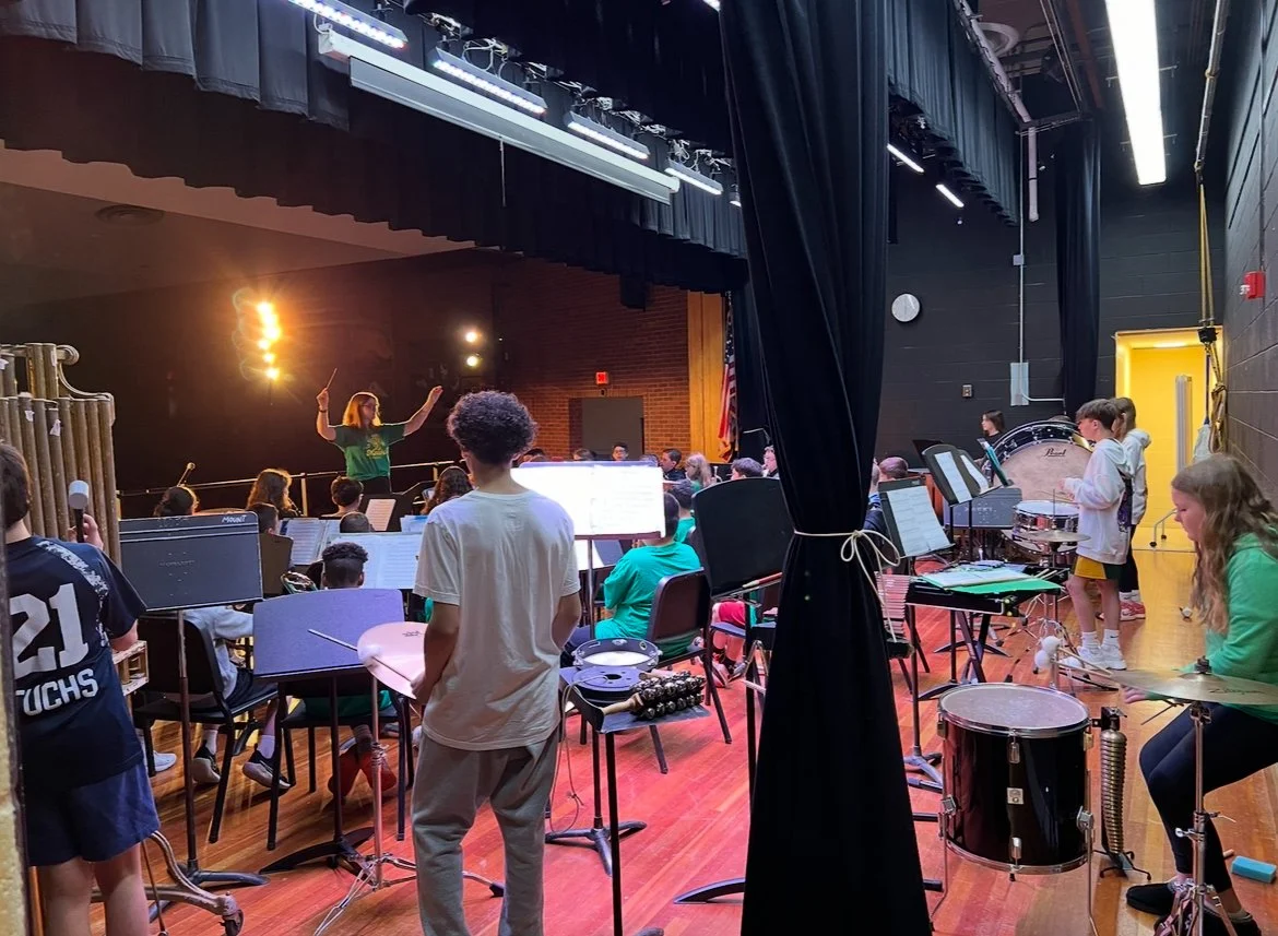 School orchestra rehearsal in a stage cafeteria with students playing various instruments, conductor leading, and music stands.