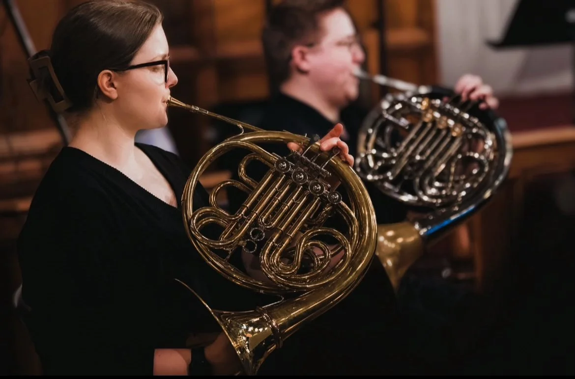 Two musicians playing French horns in an indoor setting, possibly a music room or studio.