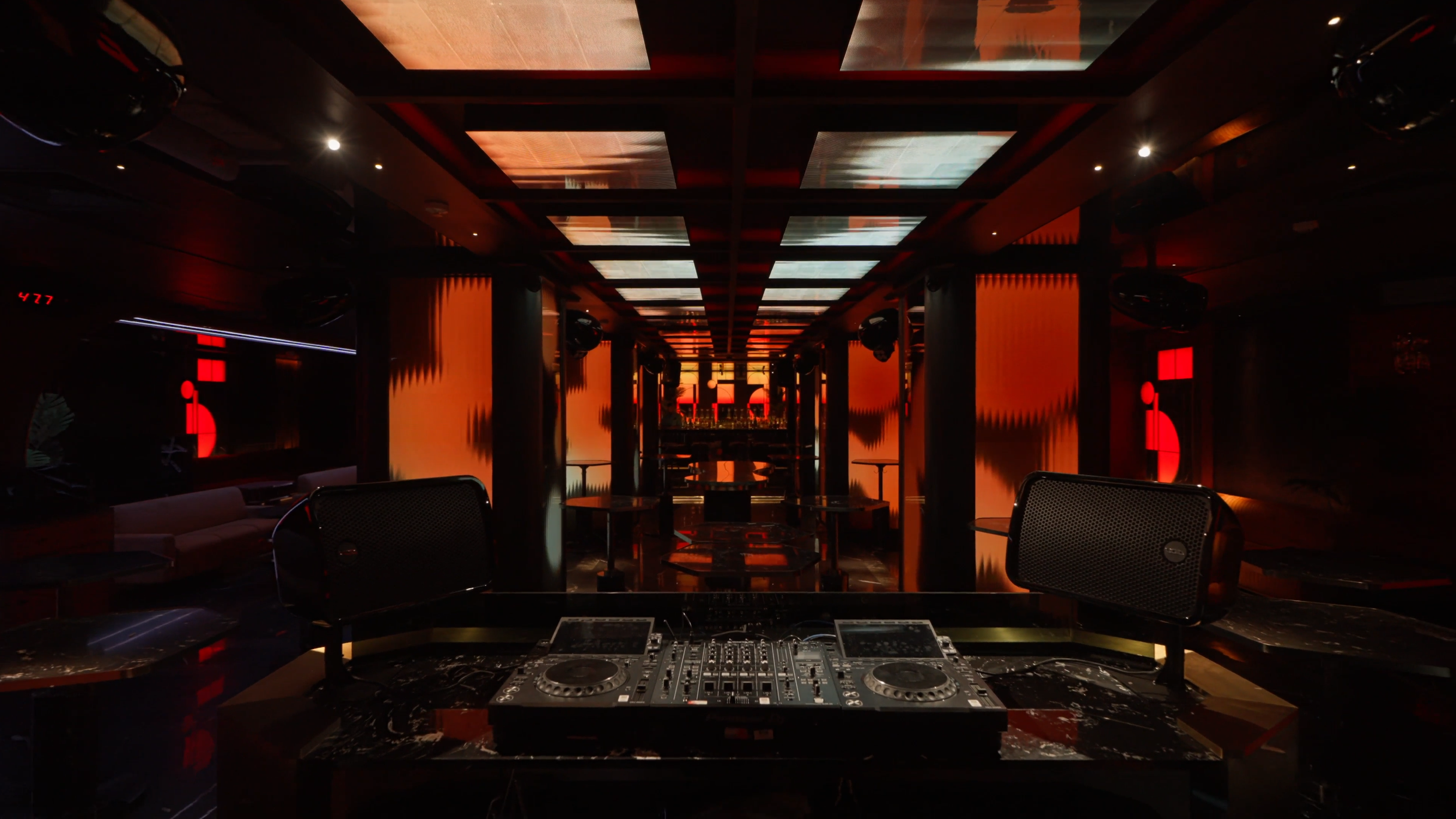 A dimly lit nightclub or lounge with a DJ setup in the foreground and a stage with red lighting and visual effects on the walls and ceiling.