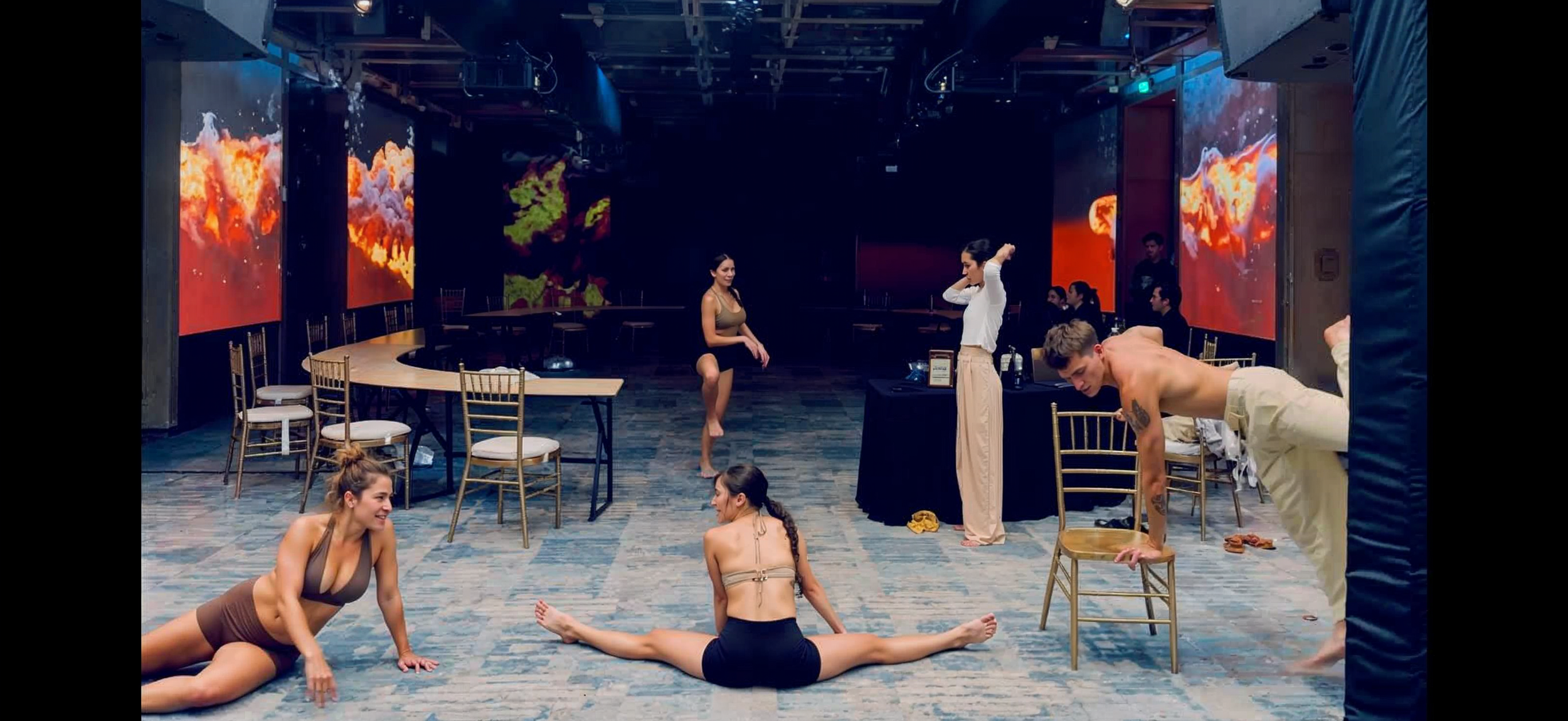 A group of people rehearsing a dance or theater performance in a studio with large screens displaying abstract fiery images in the background. Some individuals are practicing poses or stretches on the floor, while others are standing or adjusting their positions.