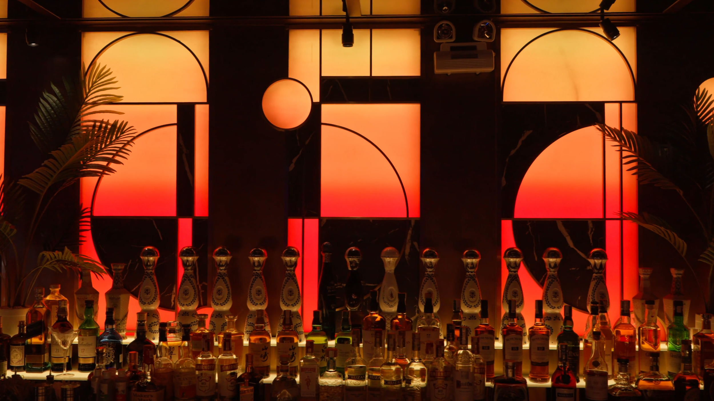 Bar with illuminated orange and yellow backlit decorative panels, shelves with various liquor bottles, and decorative plants on the sides.