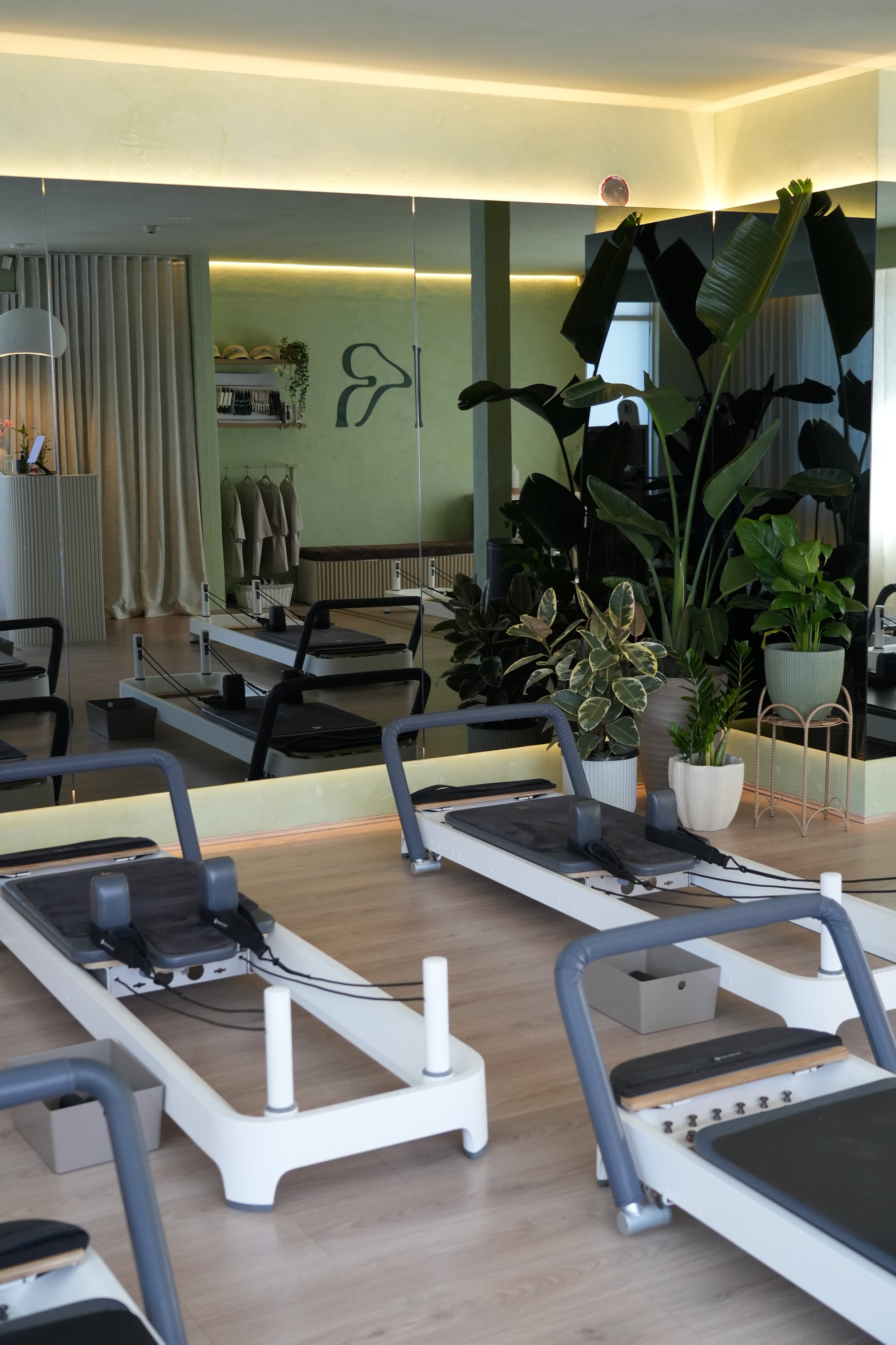 Interior view of a Pilates or yoga studio with reformer machines, large green plants, and a mirror reflecting a wall with clothing racks and decorative wall art.