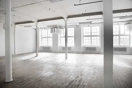 Empty industrial-style room with large windows, white walls, and wooden floors.