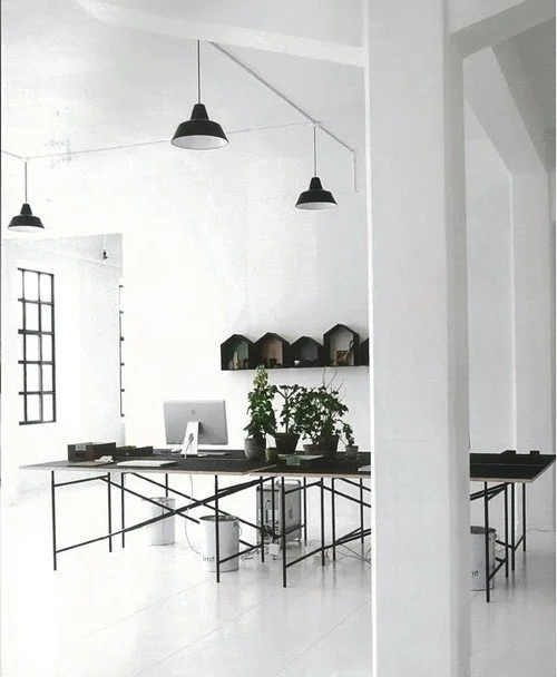 Modern, minimalist office with white walls, black pendant lights, a black table with plants, and black wall shelves with decorative items.