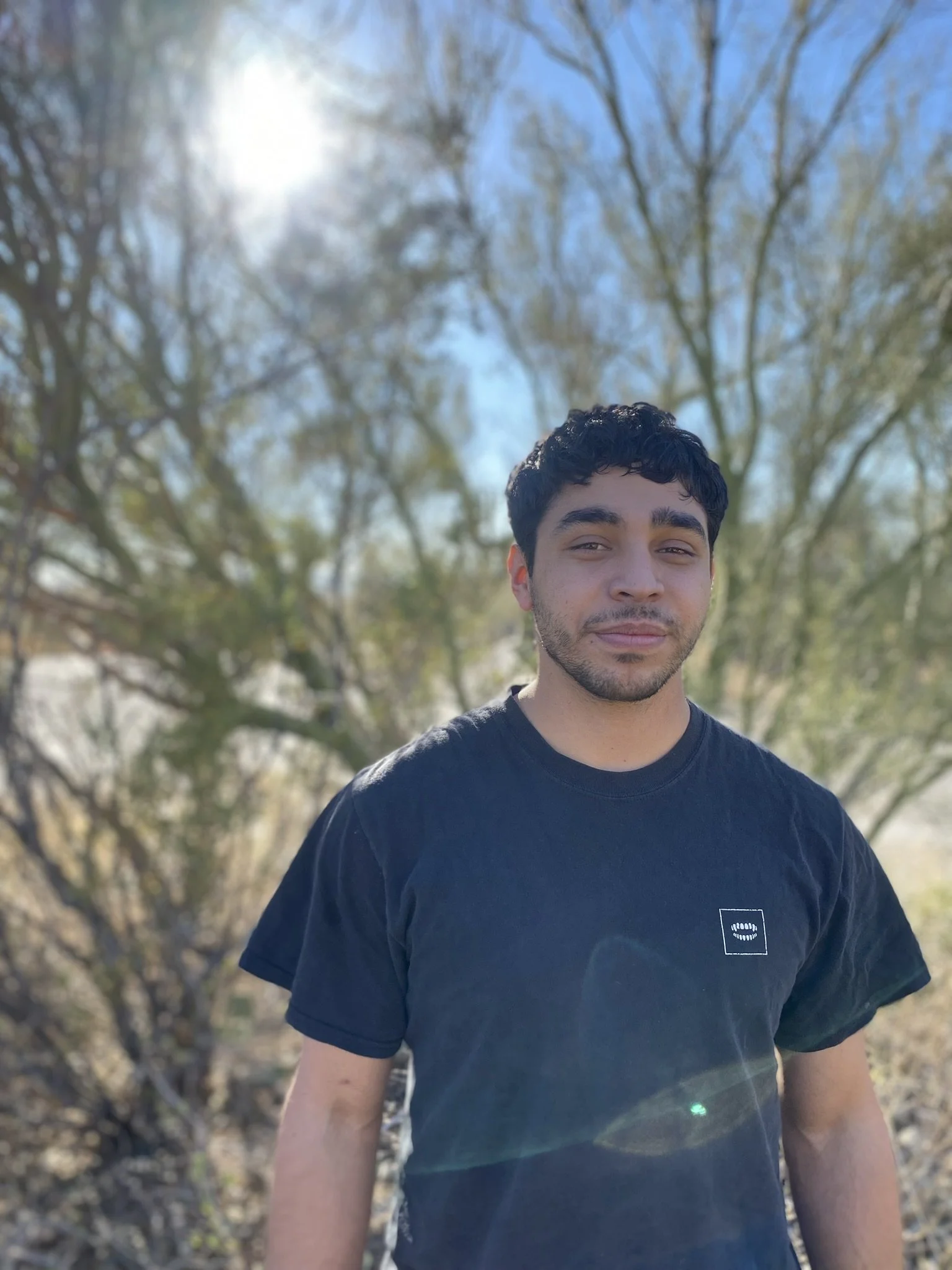 A young man with dark curly hair and light facial hair stands outside on a sunny day with a background of trees and a clear sky.