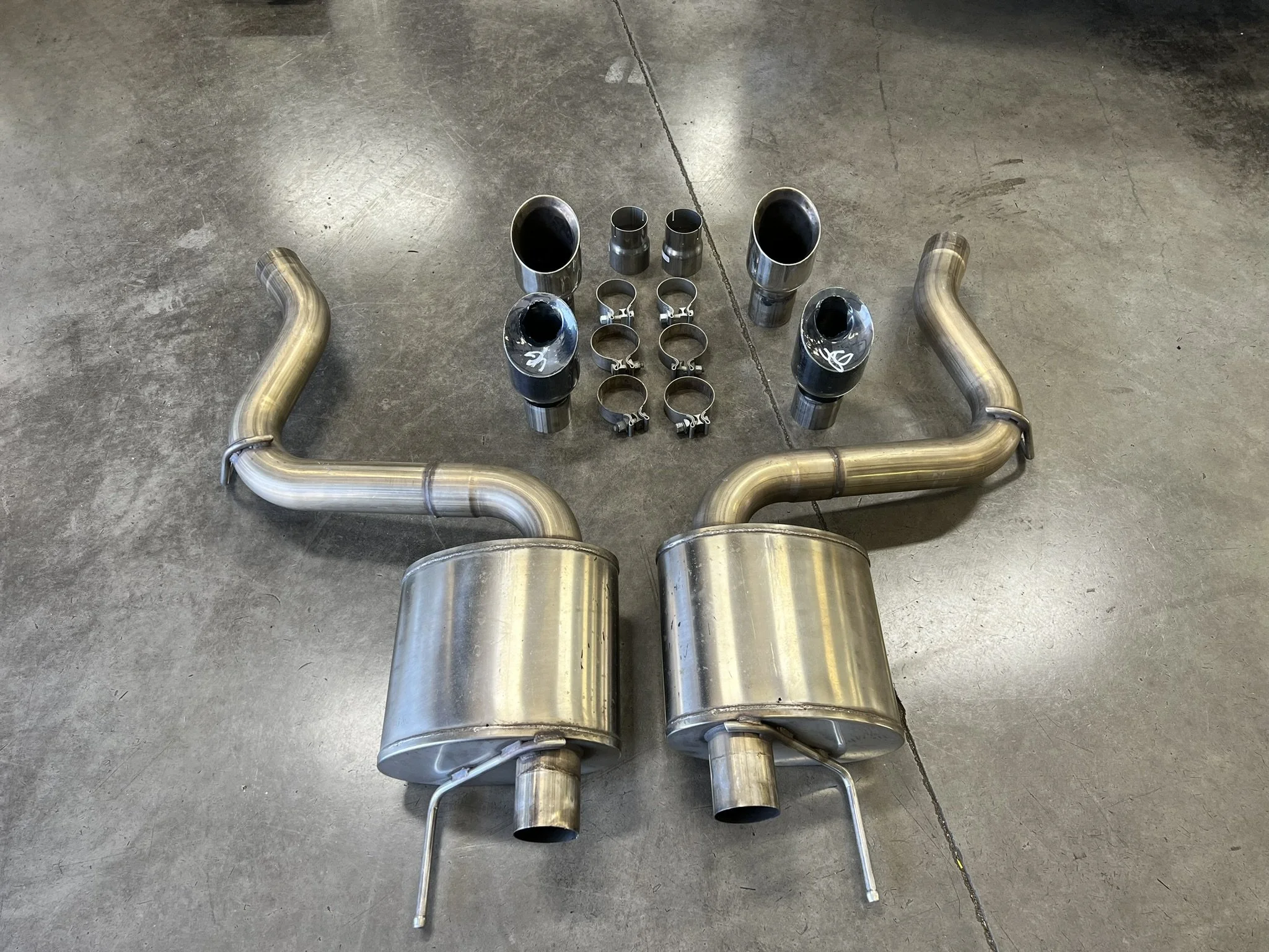 Stainless steel exhaust system components arranged on a concrete floor, including two mufflers, pipes, clamps, and connectors.