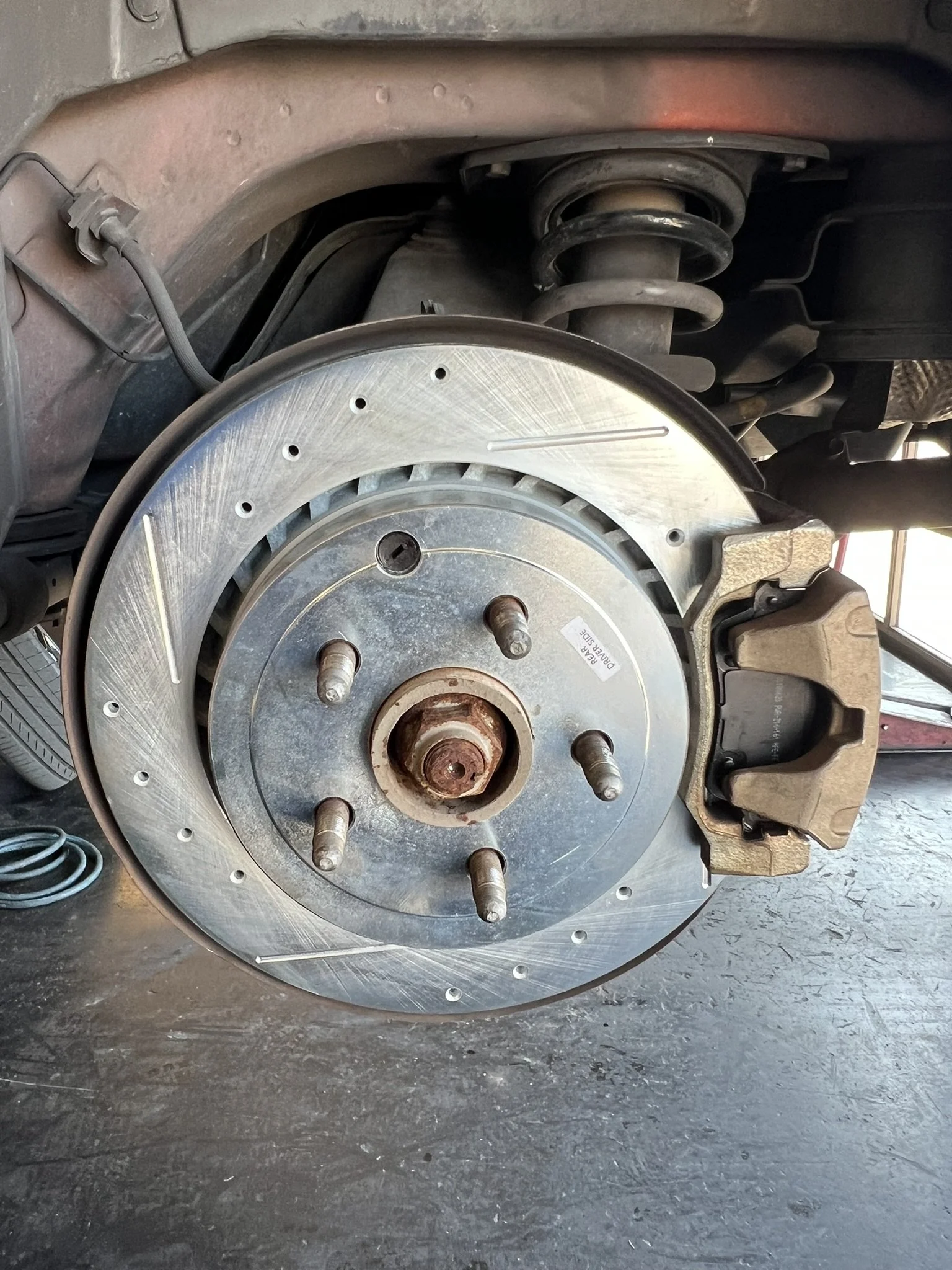 A close-up of a car's brake disc, rotor, and caliper assembly, with the wheel removed, showing the suspension components in the background.