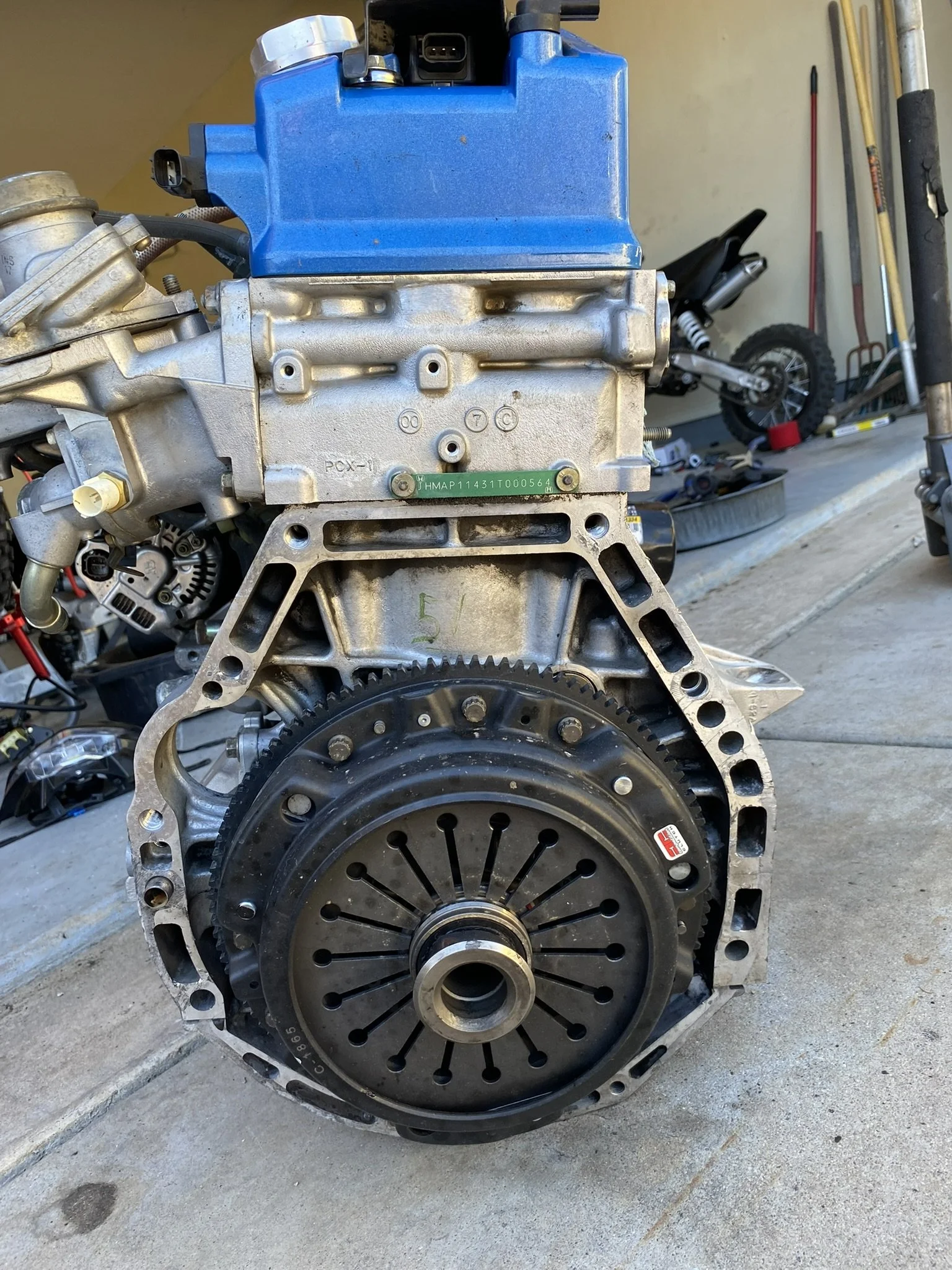Automobile engine with a blue valve cover and black clutch assembly, situated on concrete ground in front of a garage wall with tools and a bicycle in the background.