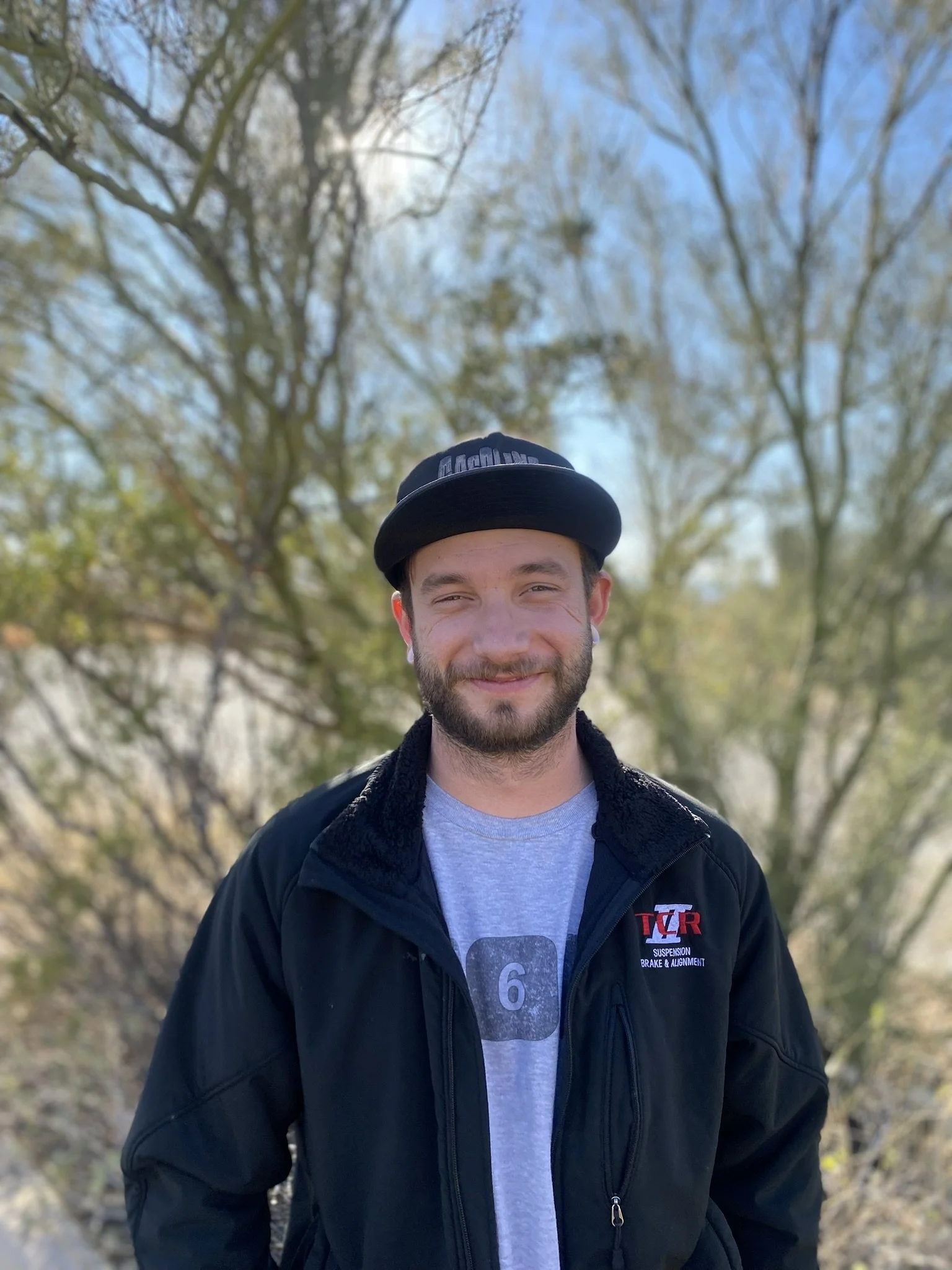 Smiling young man wearing a black baseball cap, black jacket with a logo, and a gray shirt in an outdoor setting with trees and clear sky in the background.