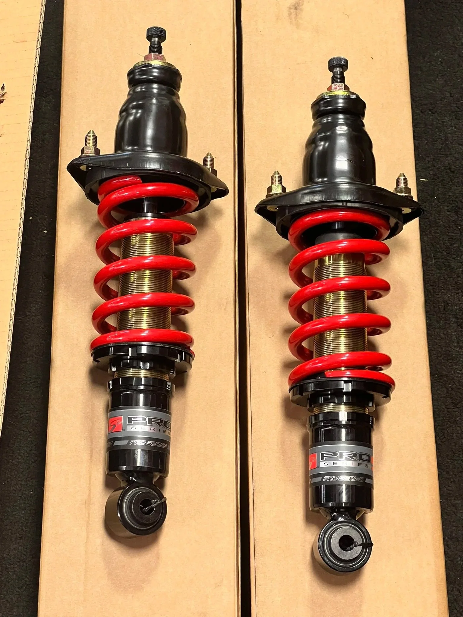 Two automotive shock absorbers with red springs and black bodies placed on cardboard boxes.