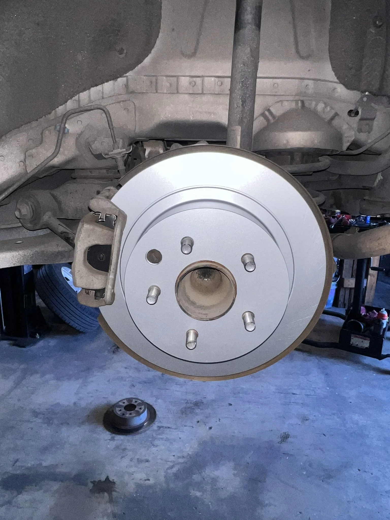 Close-up of a car's brake disc and brake caliper with missing wheel, showing multiple lug nut holes, on a concrete floor at a garage or workshop.