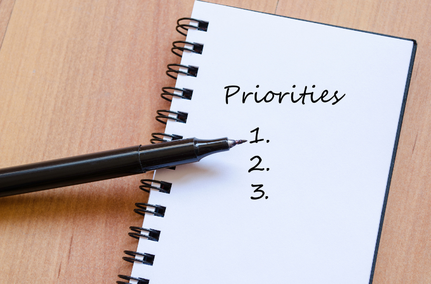 A spiral notebook on a wooden desk with the word "Priorities" written at the top and three numbered lines below for listing priorities, with a black marker placed across the notebook.