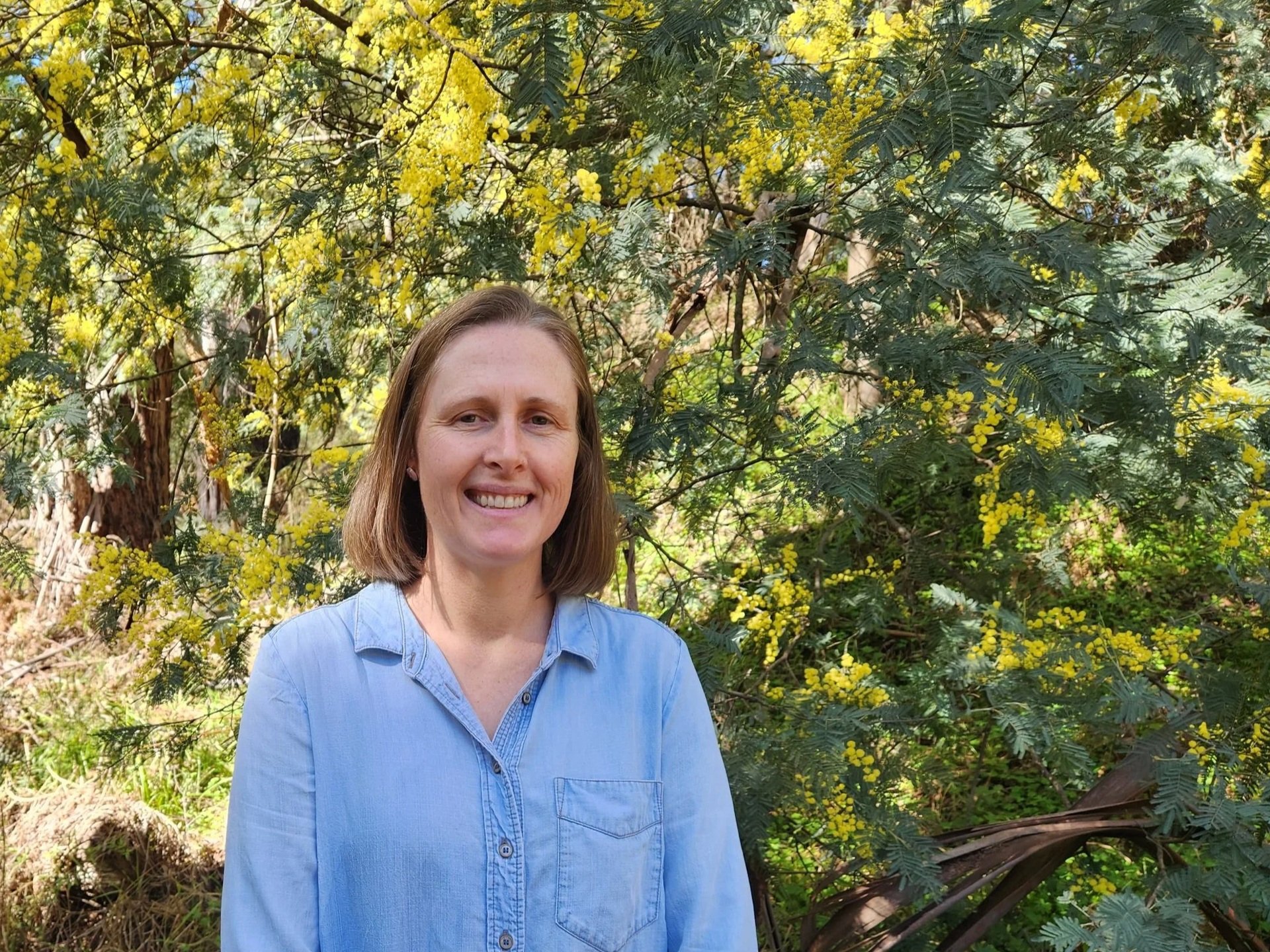 A smiling woman with shoulder-length brown hair, wearing a light blue denim shirt, standing outdoors in front of a tree with yellow flowers and green leaves.