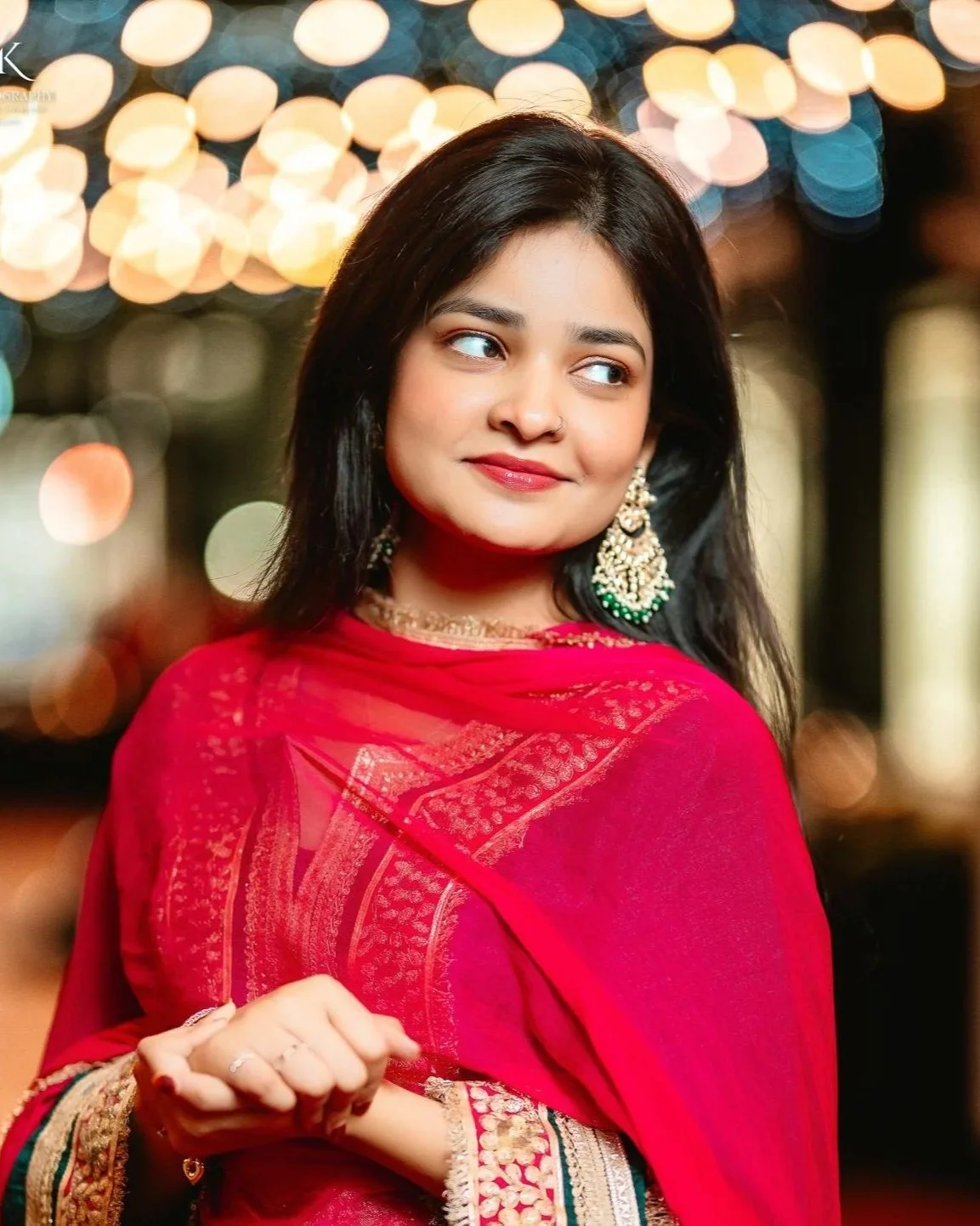 A young woman dressed in traditional Indian attire, wearing a red saree with intricate golden embroidery and jewelry, standing with her arms crossed, looking to the side with a smile, against a blurred background of lights and decor.