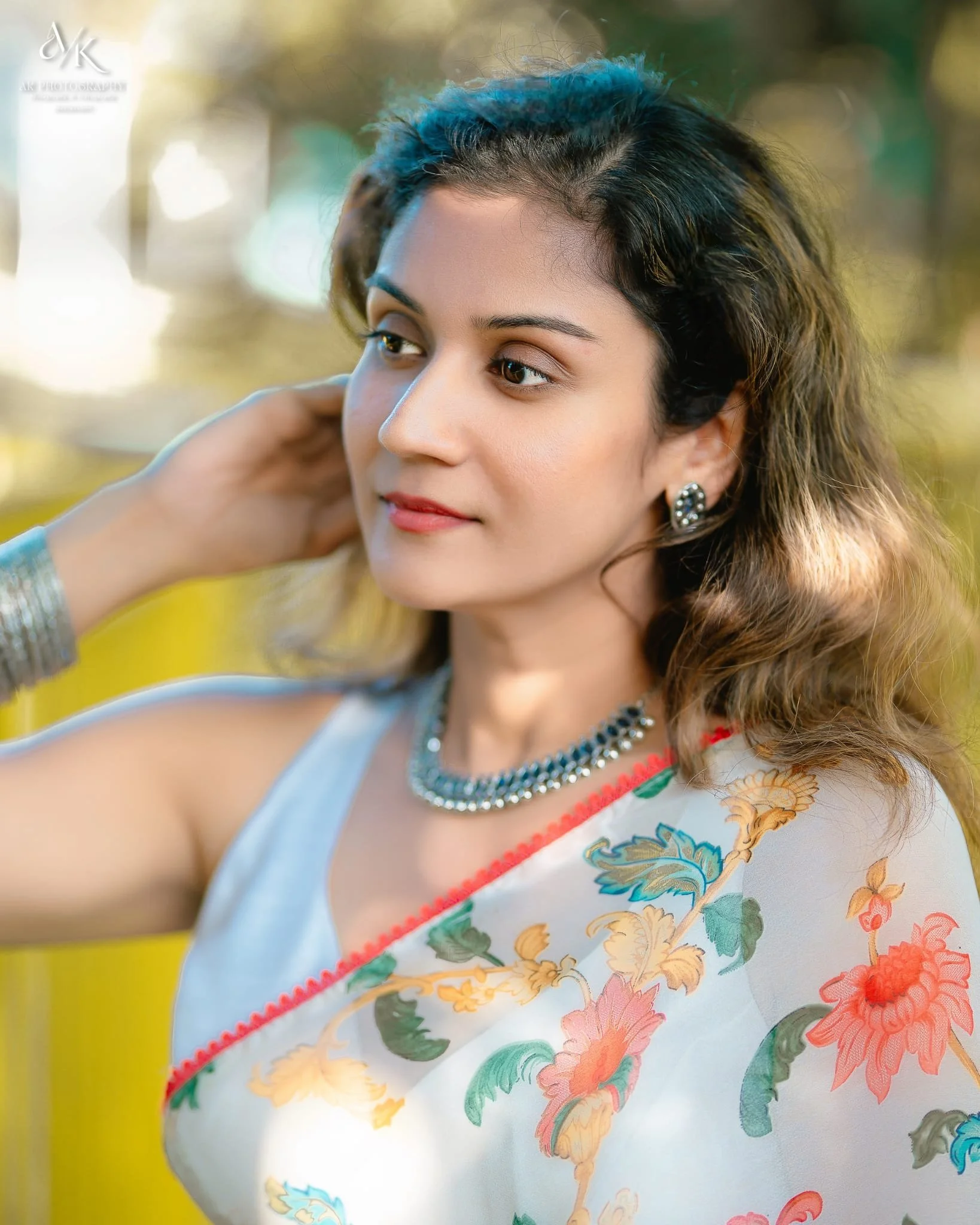 Close-up of a young woman with wavy hair wearing jewelry and a floral sari, outdoors with blurred greenery background.