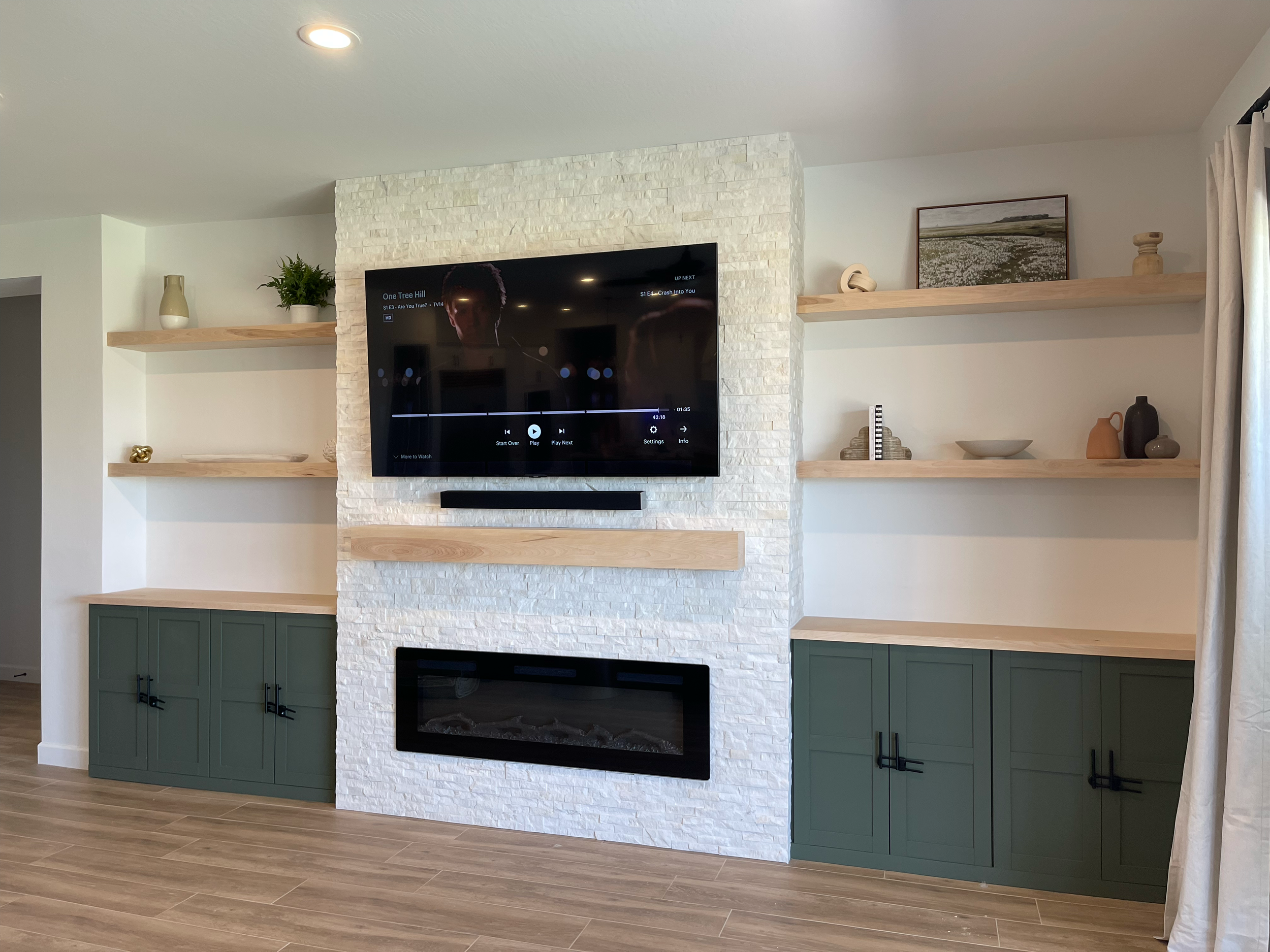 Living room with a wall-mounted TV, a fireplace below it, and built-in shelves with decorative items on a white brick wall. There are green cabinet bases and light wooden shelves.