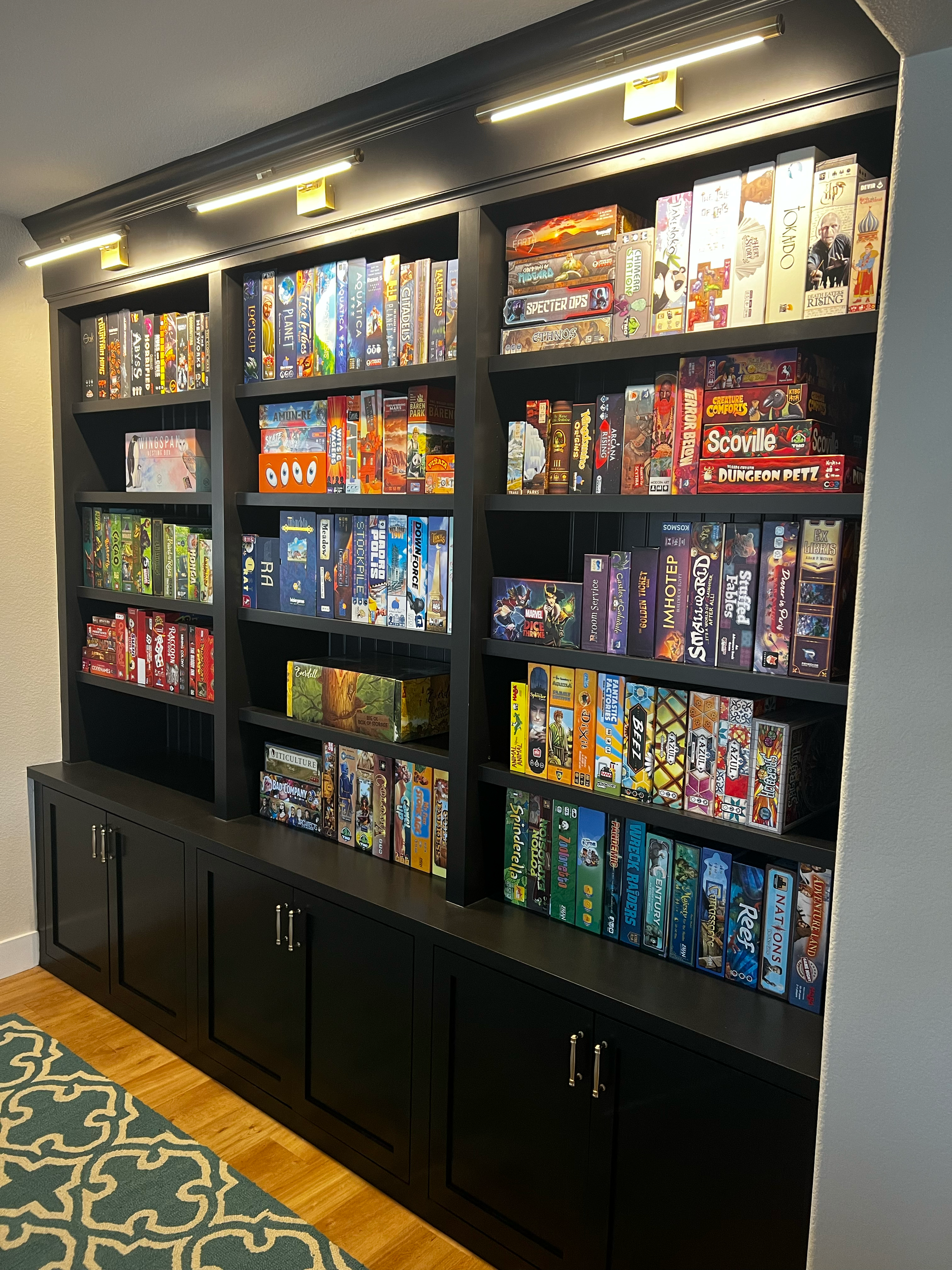 Black bookshelf filled with various board game boxes, with light fixtures above, situated in a room with a blue wall and hardwood floor.