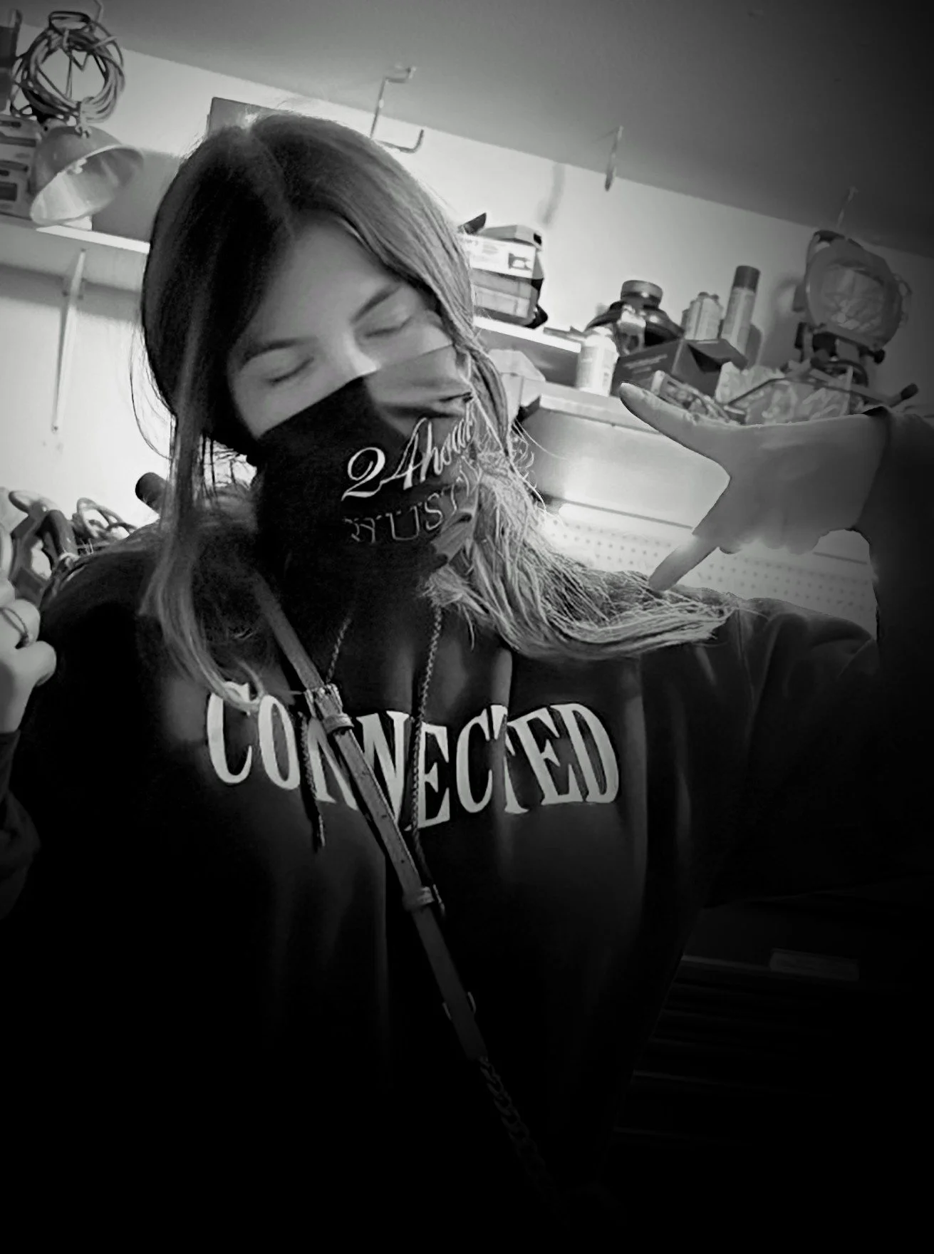 A woman wearing a face mask with 'Thanks' and 'USA' on it, a hoodie with 'CONNECTED' printed on it, making a peace sign with her hand, in a workshop or garage setting.