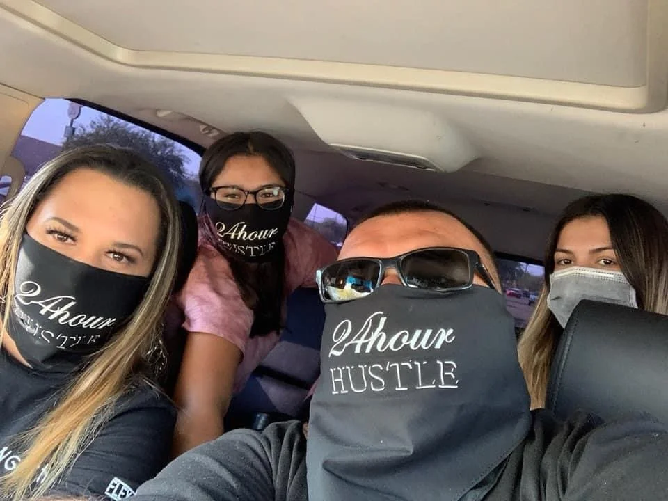 Four people sitting in a car, wearing black face masks with white text, with two women on the left and one woman on the right. The man in the center has sunglasses and a black bandana covering his lower face. The background shows a window with trees outside.