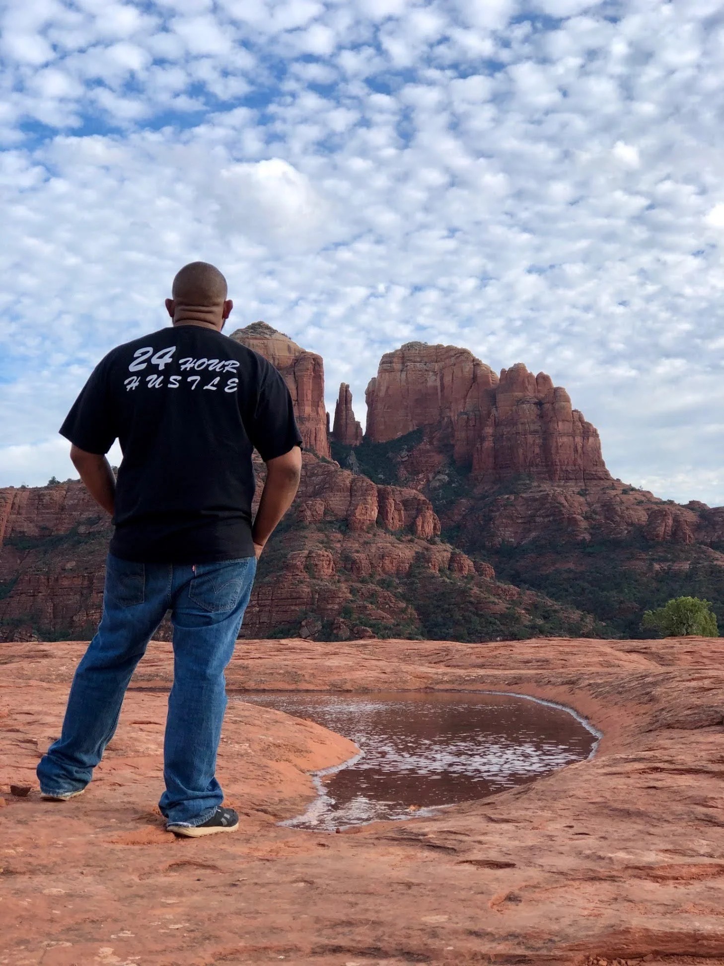A man stands on red rock in a desert landscape, looking at tall red rock formations and mountains under a sky filled with scattered clouds.