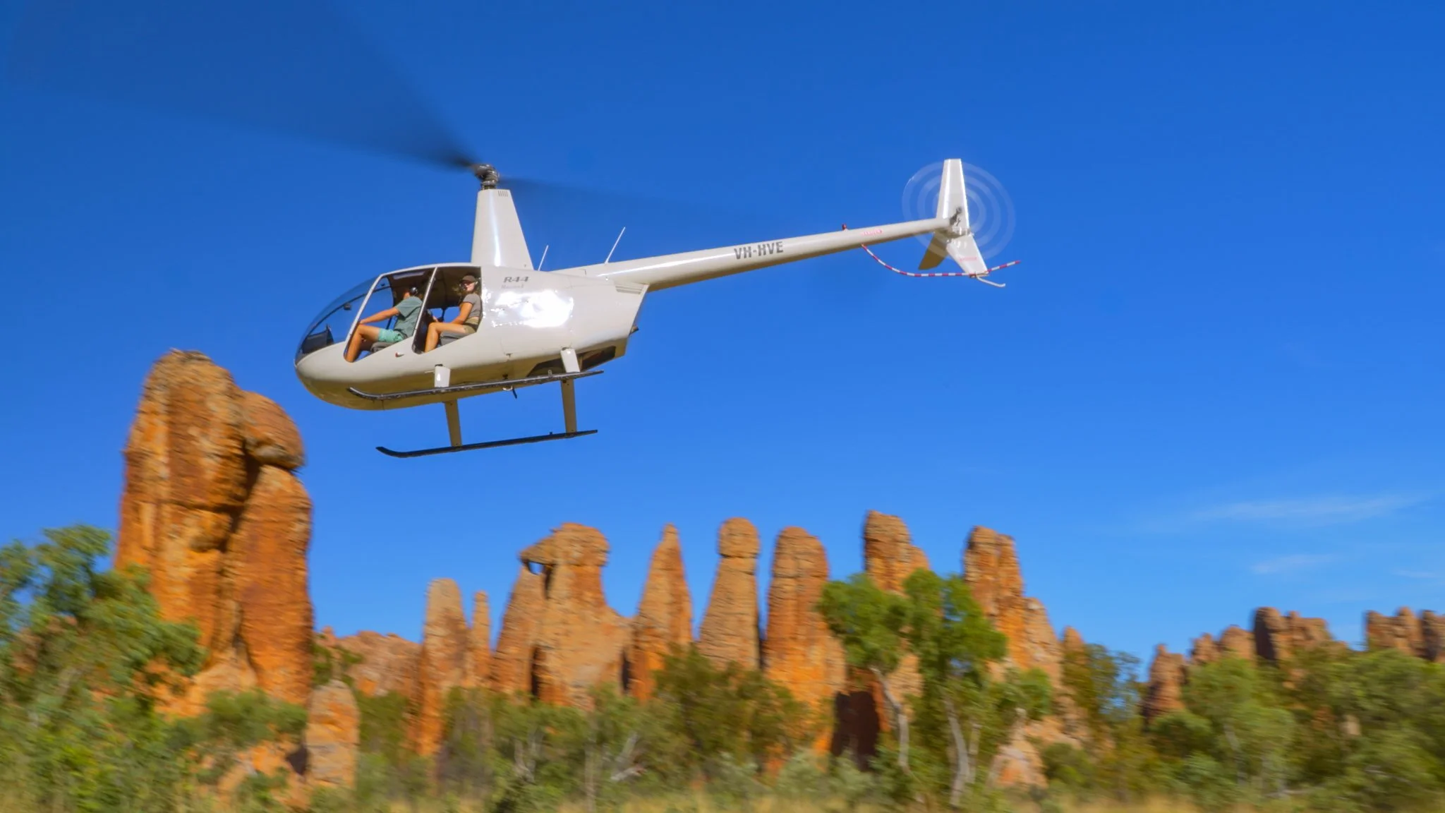 Cape Crawford, The Lost City, Northern Territory, Tour Experience. , A white helicopter flying over a landscape of red rock formations and green trees under a blue sky.