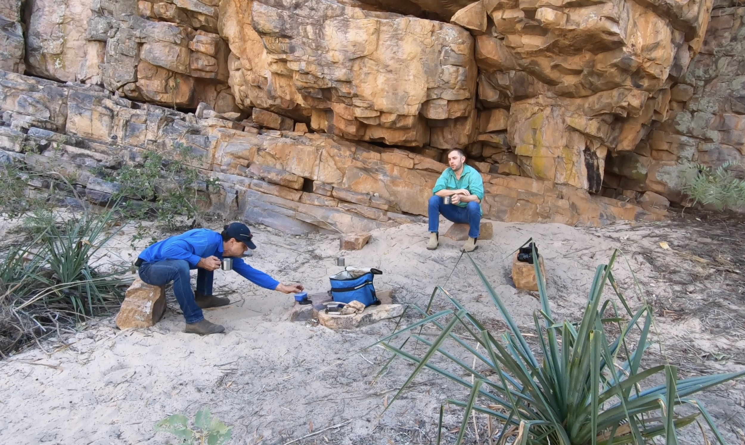 Two men sitting on rocks with camping gear in front of a rocky cliff in a natural outdoor setting, one man in a blue jacket crouching and examining a small object, the other man in a turquoise jacket sitting on a large rock holding a cup.