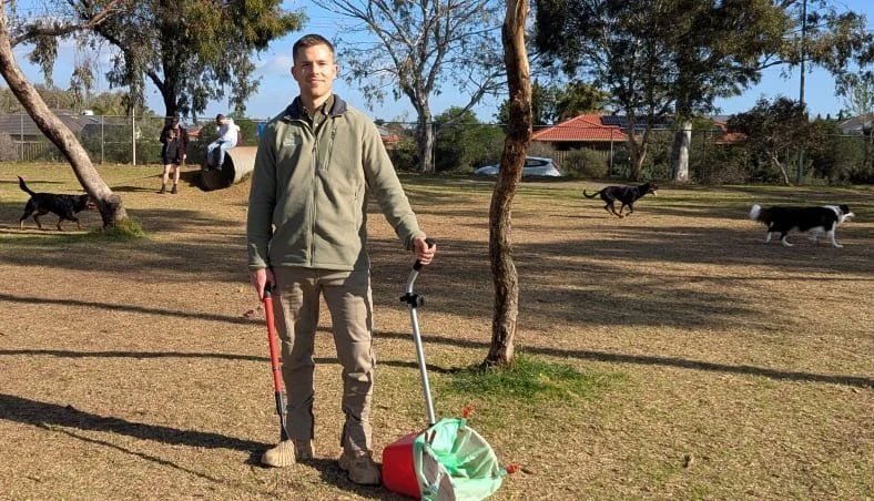 A man in uniform standing outdoors in a dog park holding a pooper scooper for dog waste removal with a garbage bag, with dogs and trees in the background.