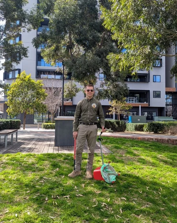 A man in uniform standing on a grassy lawn in a reserve holding a pooper scooper on the ground in front of him, behind trees and modern apartment buildings. He is ready to undertake Dog Waste Removal.