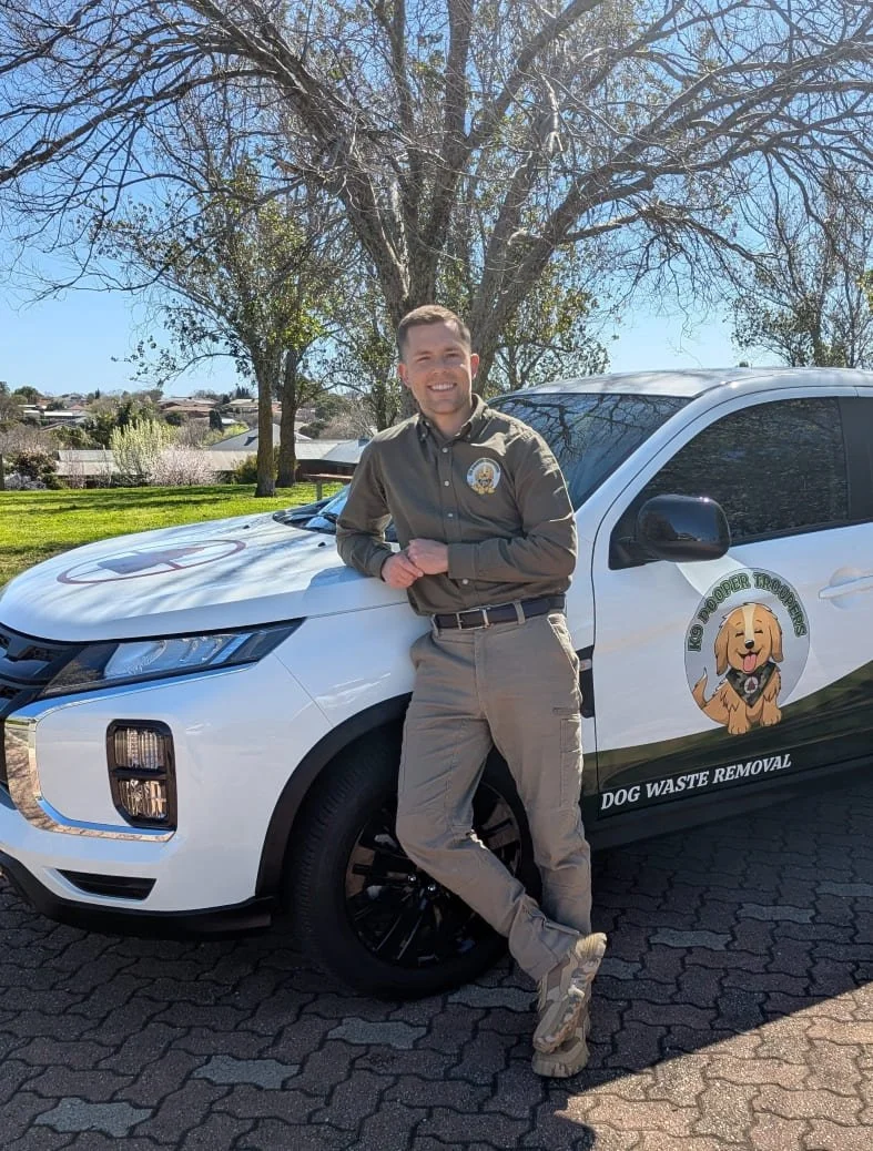 A smiling man in uniform stands besides a white Dog Waste Removal (pooper scooper) vehicle, parked on a paved area in a park with trees and houses in the background. The vehicle has graphics of the K9 Pooper Troopers logo and company information.