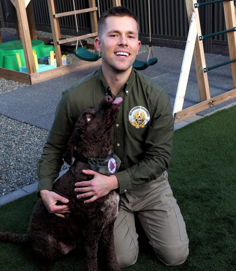 A smiling man in uniform from K9 Pooper Troopers kneeling on grass with a brown, curly-coated dog attempting to lick him in an outdoor play area with swings and play structures.