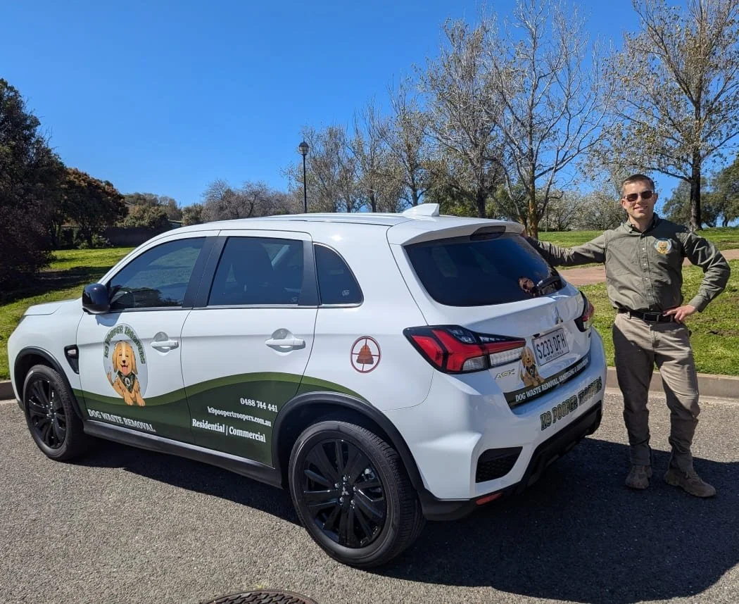 A smiling man in a uniform standing next to a white K9 Pooper Troopers dog waste removal vehicle parked on a road next to a park with green grass and trees. The vehicle has graphics of their logo, which features a cute dog, and the business details.