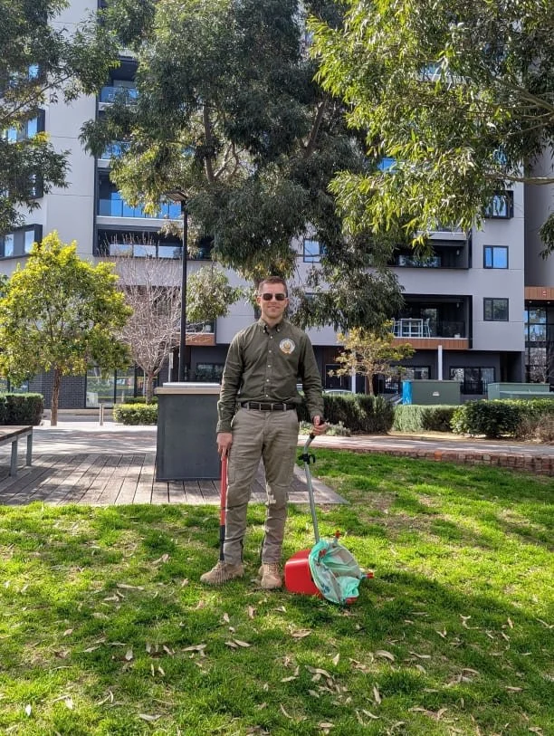 A man in uniform standing on a grassy lawn in a reserve holding a pooper scooper on the ground in front of him, behind trees and modern apartment buildings. He is ready to undertake Dog Waste Removal.