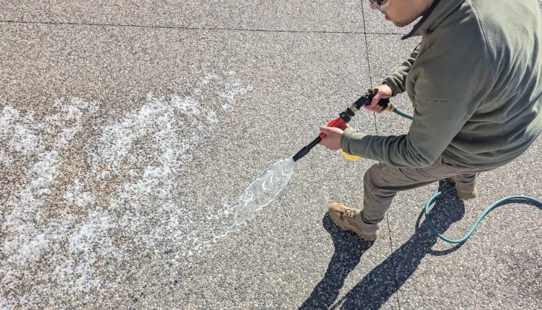 A man using a hose spray gun to clean (sanitise and deodorise) an aggregate concrete surface, with cleaning solution spraying from the nozzle.