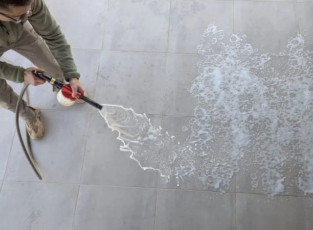 A man using a hose spray gun to sanitise and deodorise (clean) a tiled floor, with chemical suds spread across the surface.