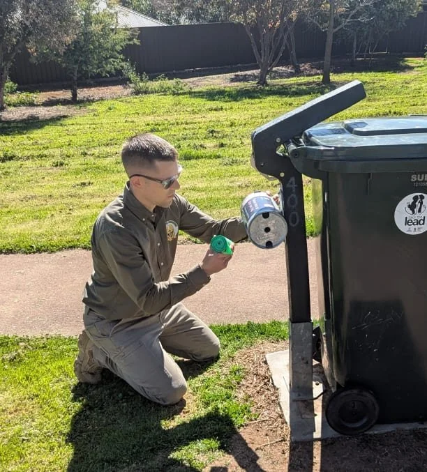 A man from K9 Pooper Troopers in uniform kneeling on the grass inspecting a dog waste bag dispenser (also called a dog poop bag dispenser) with bags in hand to simulate replenishment.