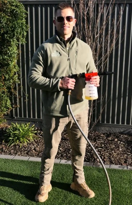 A man standing outdoors holding a hose sprayer gun for Sanitising & Deodorising, wearing sunglasses, a green jacket, khaki pants, and beige shoes.