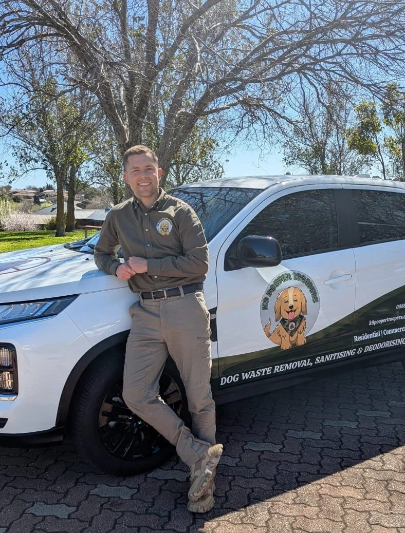 A smiling man in uniform stands besides a white Dog Waste Removal (pooper scooper) vehicle, parked on a paved area in a park with trees and houses in the background. The vehicle has graphics of the K9 Pooper Troopers logo and company information.