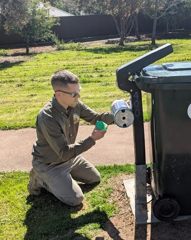 A man from K9 Pooper Troopers in uniform kneeling on the grass inspecting a dog waste bag dispenser (also called a dog poop bag dispenser) with bags in hand to simulate replenishment.