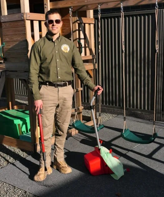 A man in sunglasses and outdoor work attire standing on a rubber mat in front of a wooden playset, holding a pooper scooper for dog poo pickup/cleanup, with swings hanging behind him.
